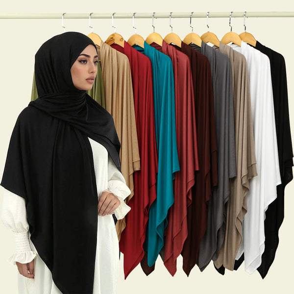 A woman wearing a black hijab and white clothing stands in front of a rack displaying various colored modal square scarves.