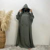 Une personne portant une abaya longue vert olive foncé avec capuche et manches batwing, debout contre un mur blanc.