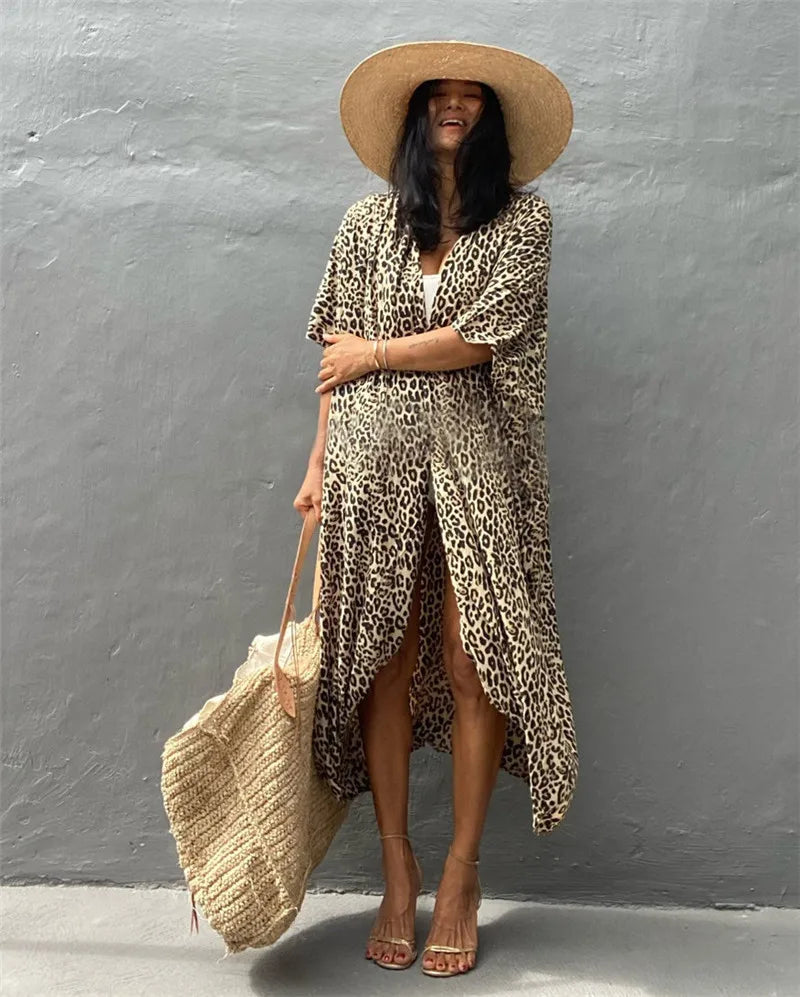 Eine Frau in einem Leopardenmuster-Maxikleid und einem breiten Strohhut lehnt vor einer grauen Wand und hält eine geflochtene