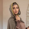 Eine Frau trägt einen beigeen Spitzen-Chiffon-Hijabschal mit goldenem Zierrand und blickt direkt in die Kamera.