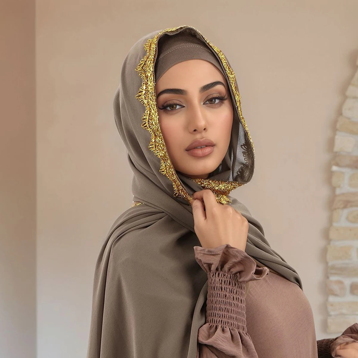 Eine Frau trägt einen beigeen Spitzen-Chiffon-Hijabschal mit goldenem Zierrand und blickt direkt in die Kamera.