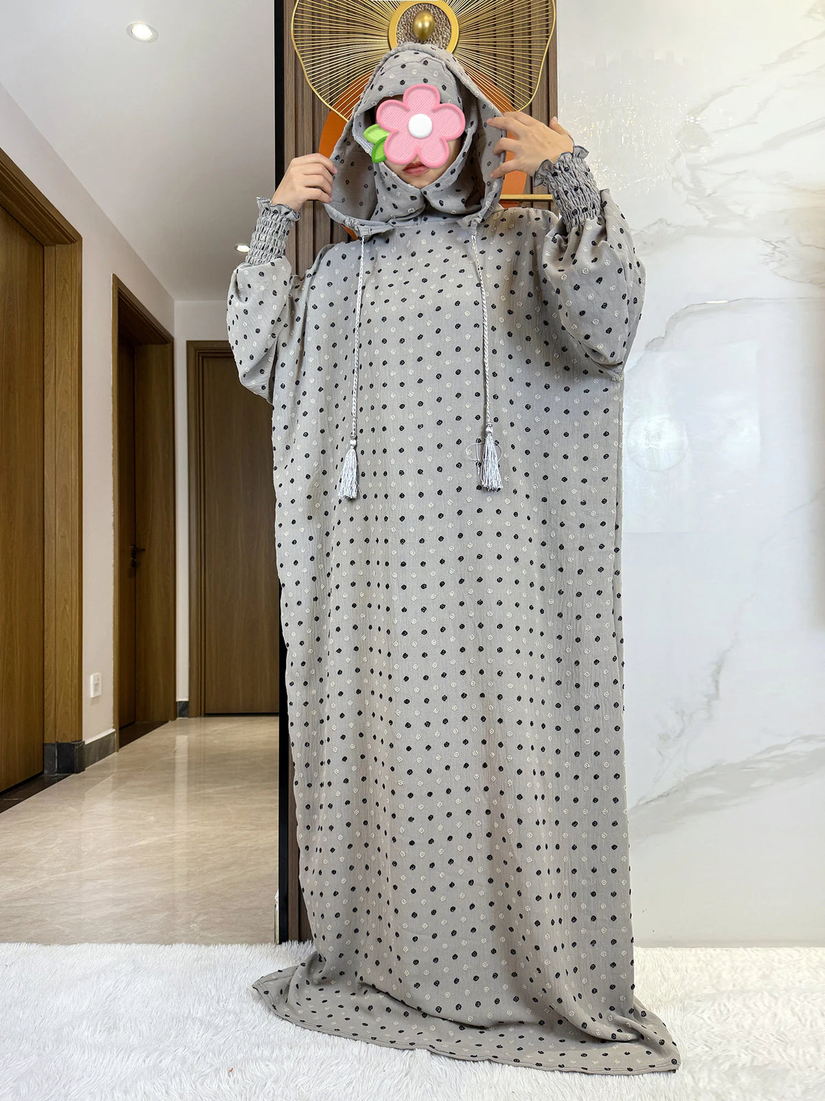 Una larga abaya de algodón gris con capucha y estampado de puntos, exhibida sobre un fondo de mármol blanco.