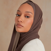Eine Frau mit braunem Chiffon-Hijab und weißem Oberteil, die direkt in die Kamera schaut.