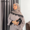 Una mujer vistiendo un conjunto de abaya de encaje beige con un hiyab a juego y sosteniendo un clutch acolchado negro.