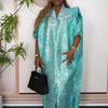 A woman wearing a teal satin kaftan abaya with gold jewelry and a matching turban, standing in front of a light pink wall.