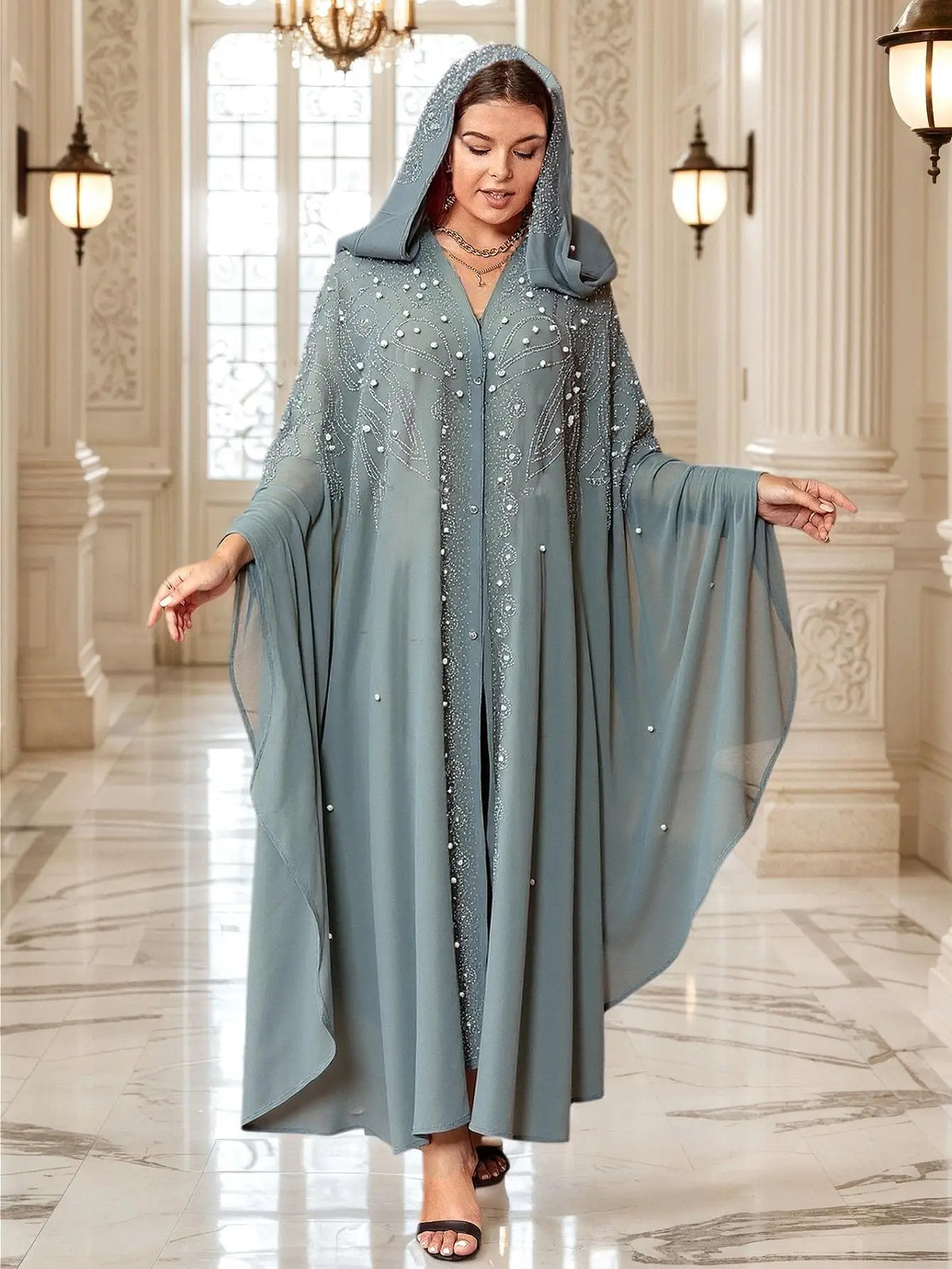 Una mujer modela un maxi vestido abaya azul polvoriento y fluído con mangas de murciélago en un pasillo bien iluminado.