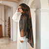 A woman wearing a sheer brown watercolor print hijab and white traditional dress stands in a bright interior hallway.
