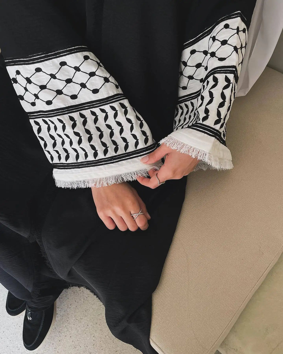 Gros plan sur une abaya noire avec broderie géométrique blanche sur les manches et le bas, portée par une personne.