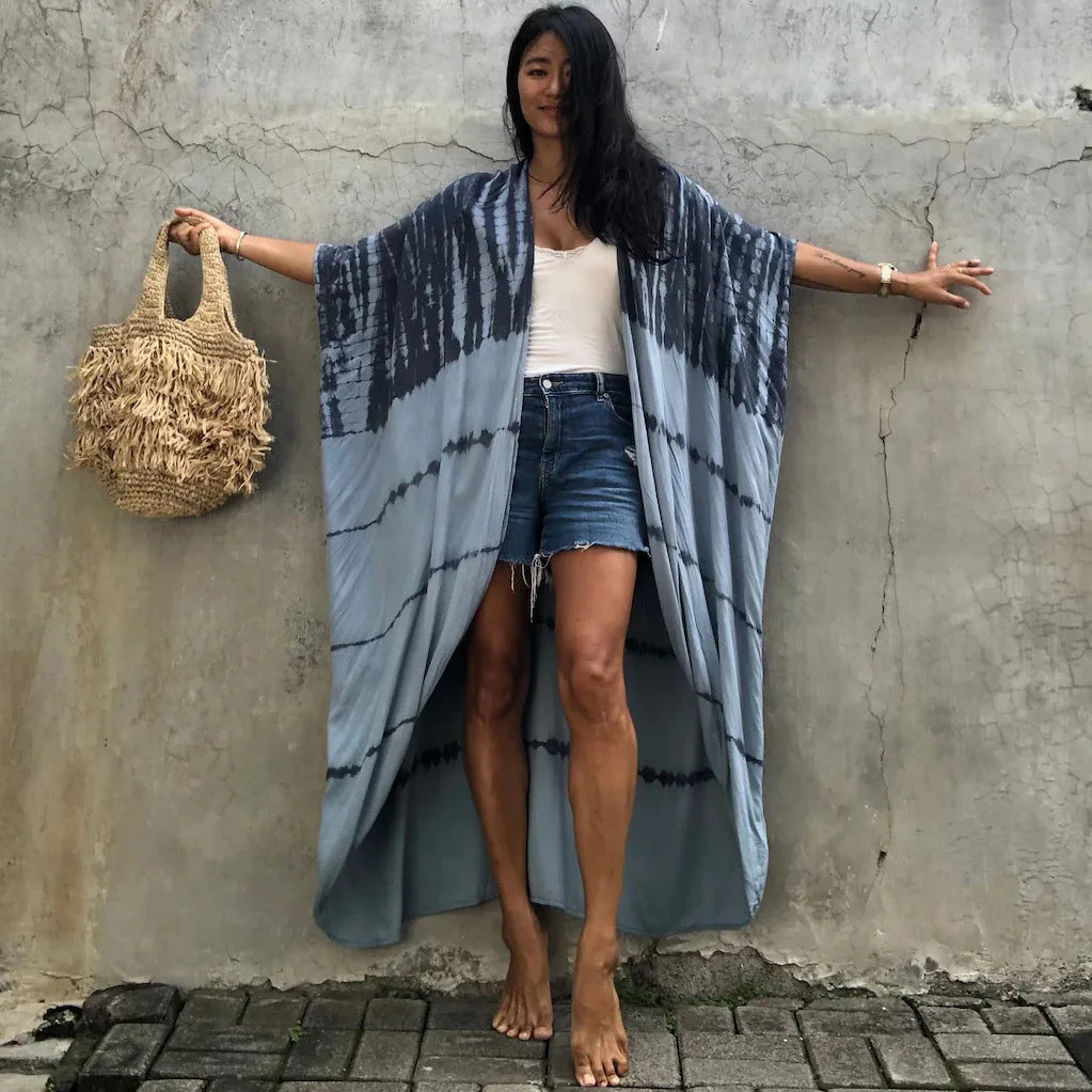 Eine Frau in einem blau-grau gemusterten Kimono-Überwurf über Jeansshorts vor einer Betonwand.