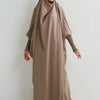 Eine Frau in einem beige abgetfitzten Abaya mit gerafften Ärmeln vor einer weißen Wand.