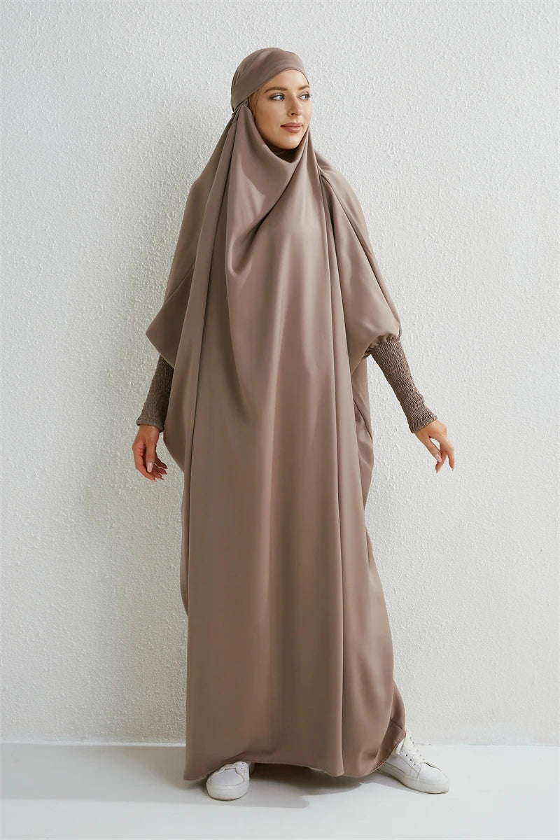 Eine Frau in einem beige abgetfitzten Abaya mit gerafften Ärmeln vor einer weißen Wand.