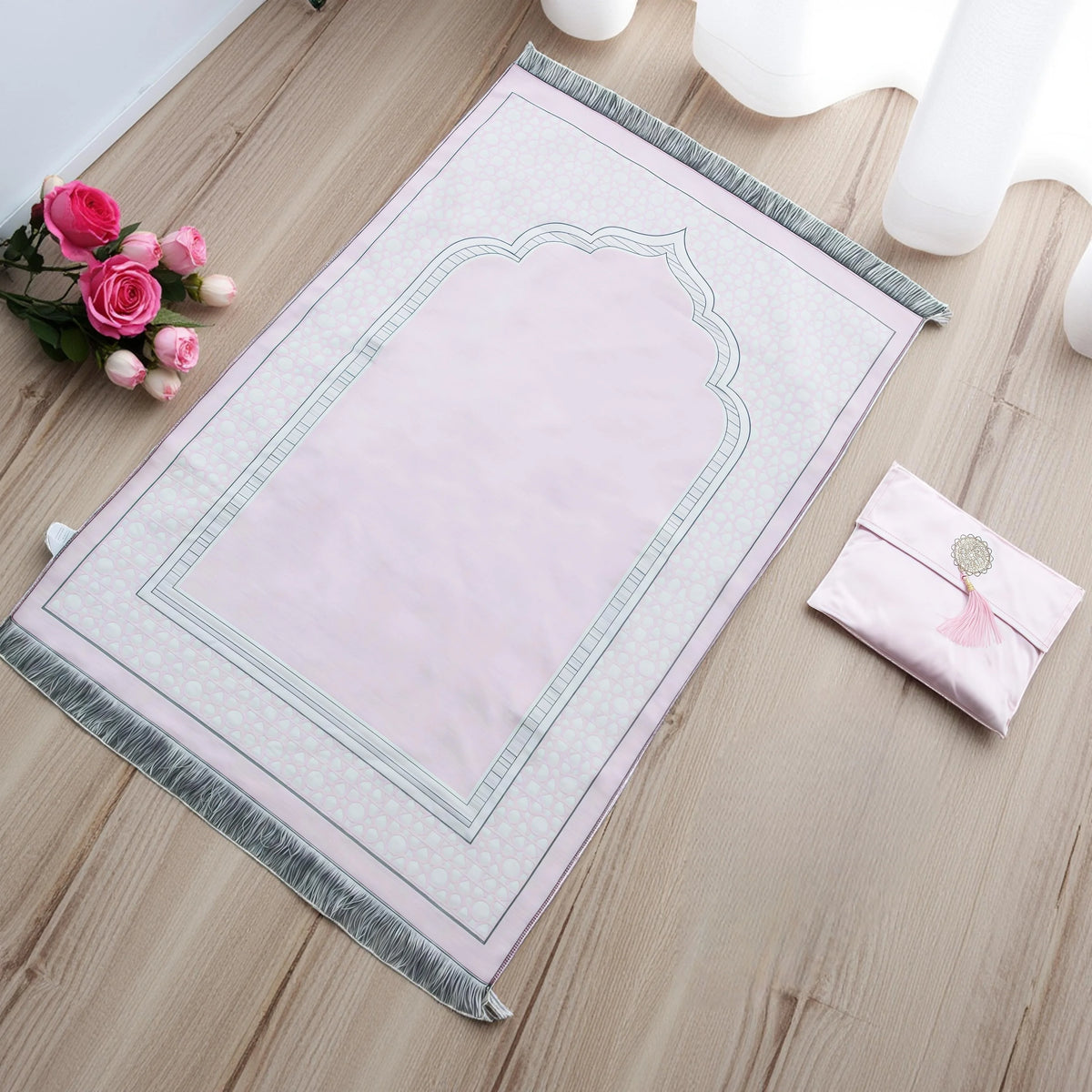 Alfombra de oración musulmana rosa con borde decorativo y una bolsa a juego, sobre un suelo de madera clara.