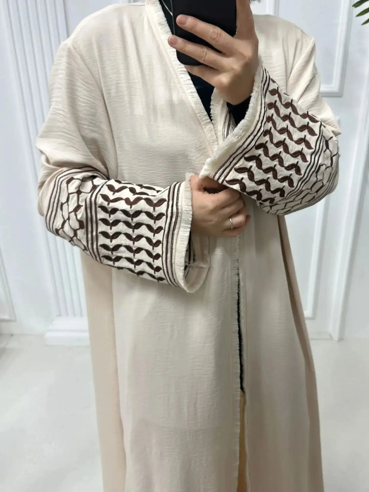 Une femme portant une élégante abaya beige à manches brodées et un haut noir, tenant un téléphone.