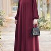 Una mujer con un vestido maxi holgado color granate y un hiyab beige sostiene un bolso negro al aire libre.