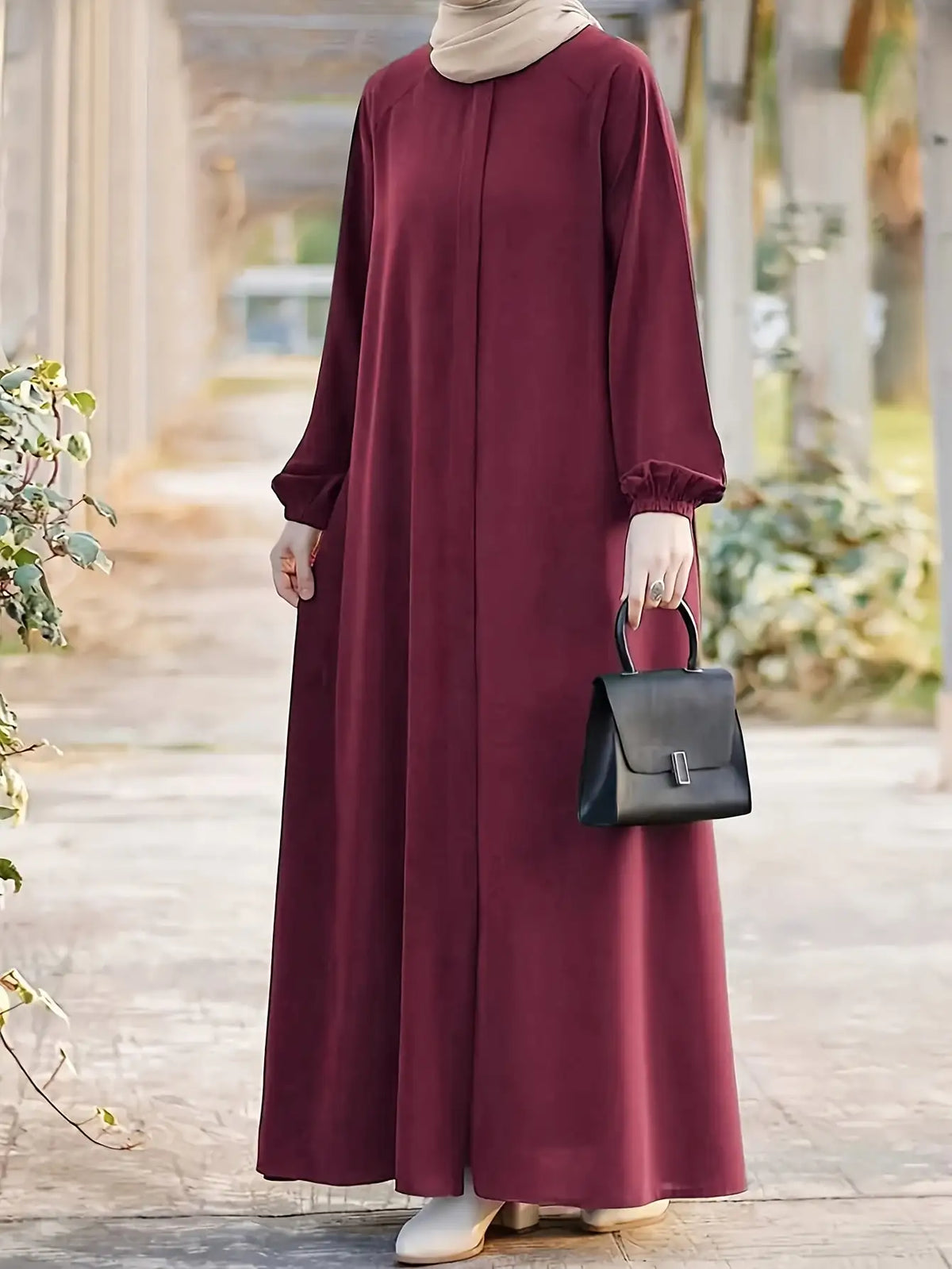 Una mujer con un vestido maxi holgado color granate y un hiyab beige sostiene un bolso negro al aire libre.