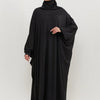 Una mujer vistiendo una elegante abaya negra con capucha y hijab, de pie contra un fondo claro.