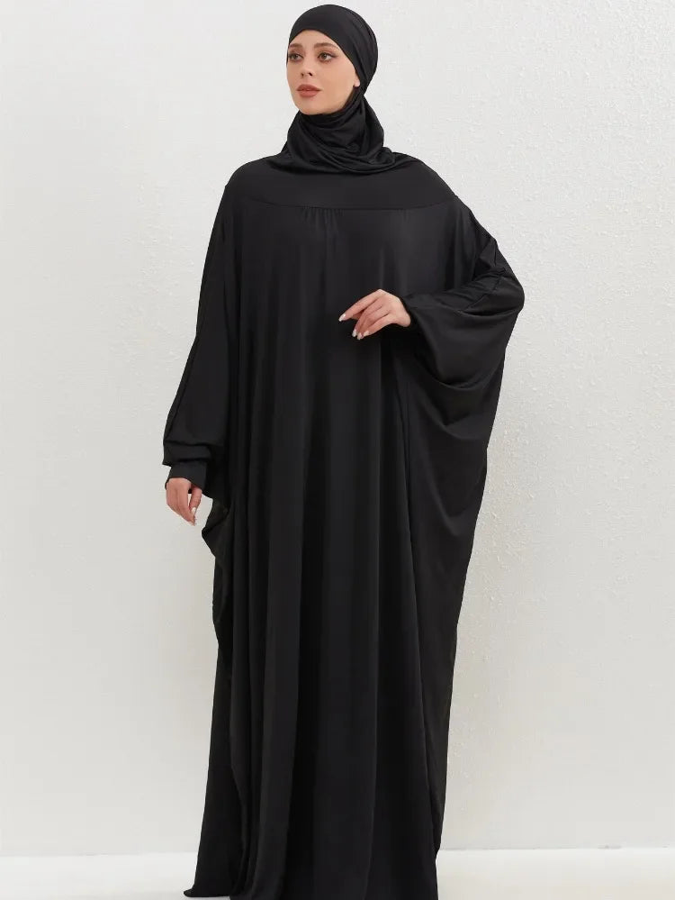 Una mujer vistiendo una elegante abaya negra con capucha y hijab, de pie contra un fondo claro.