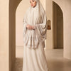 Eine Frau trägt ein elegantes beige, mehrlagiges Spitzen-Khimar-Abaya-Set in einem hellen, gewölbten Flur.