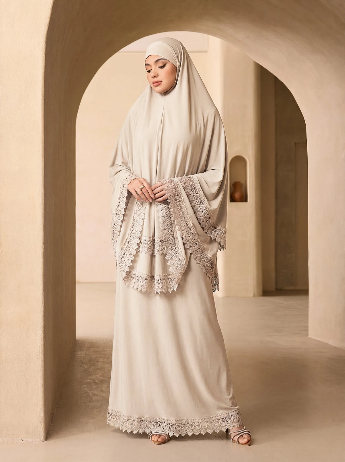 Eine Frau trägt ein elegantes beige, mehrlagiges Spitzen-Khimar-Abaya-Set in einem hellen, gewölbten Flur.