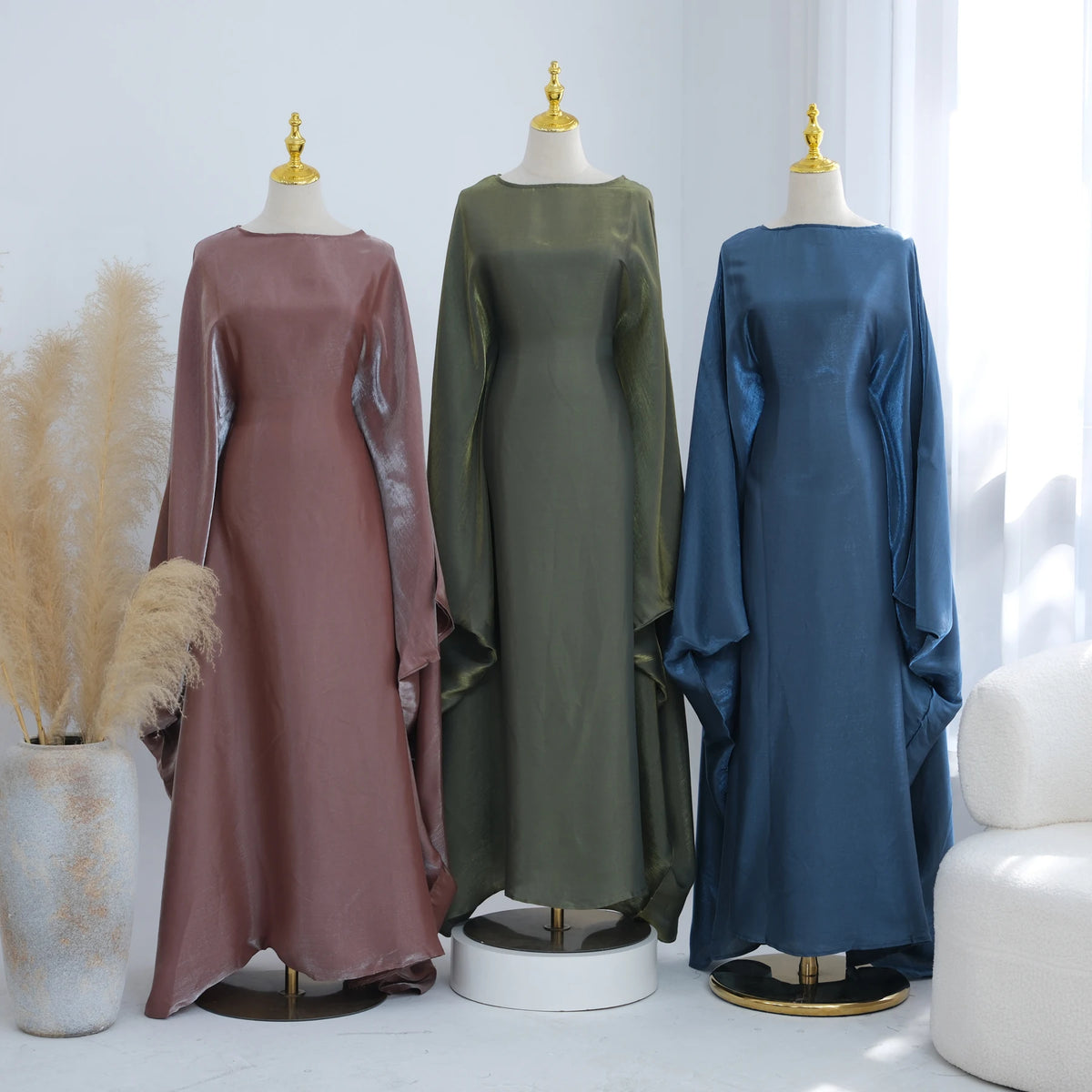 Drei elegante Kaftan-Kleider in Mauve, Olivgrün und staubigem Blau auf Mannequins.