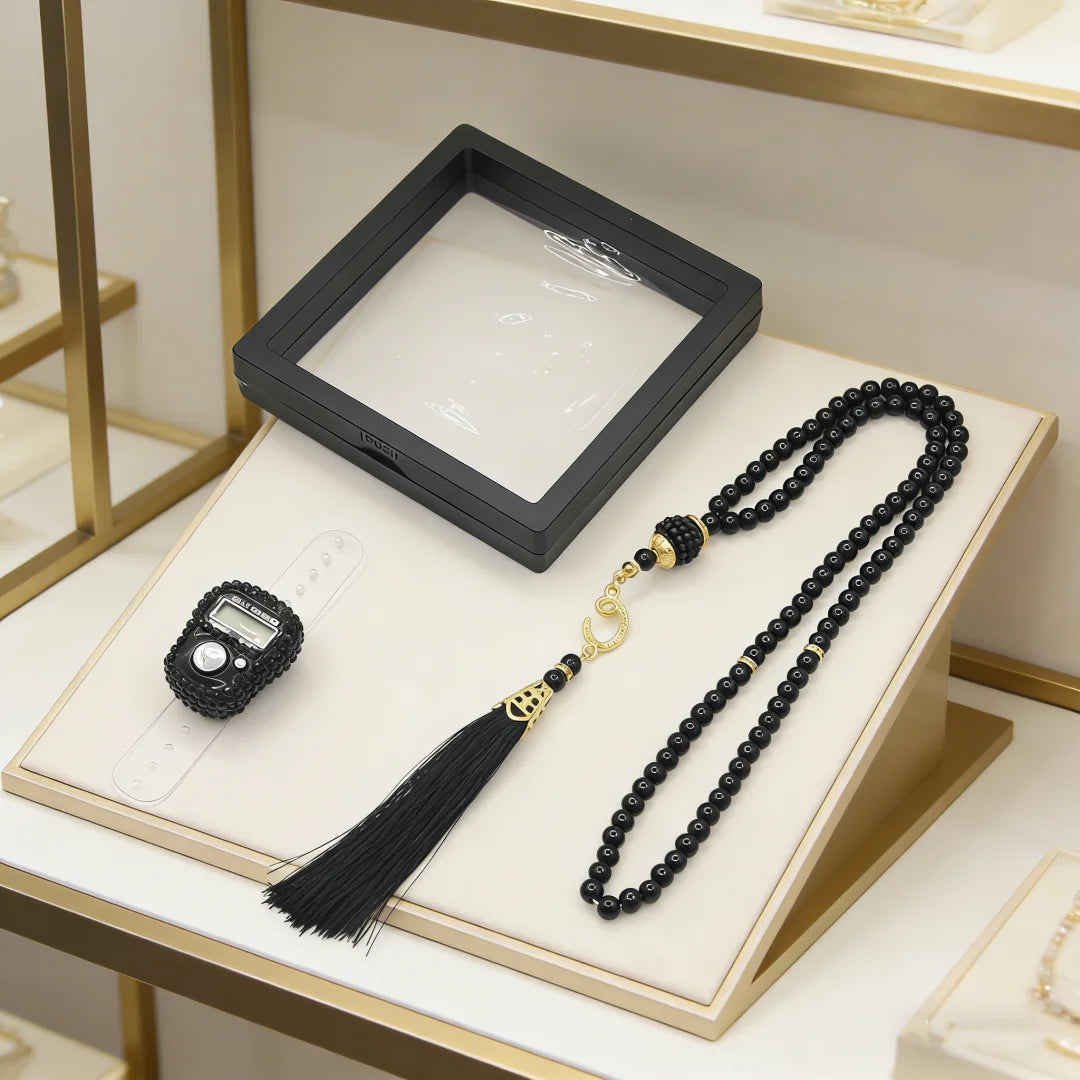 A black Islamic tasbih prayer beads with gold accents and a digital counter bracelet displayed on a light wooden tray.