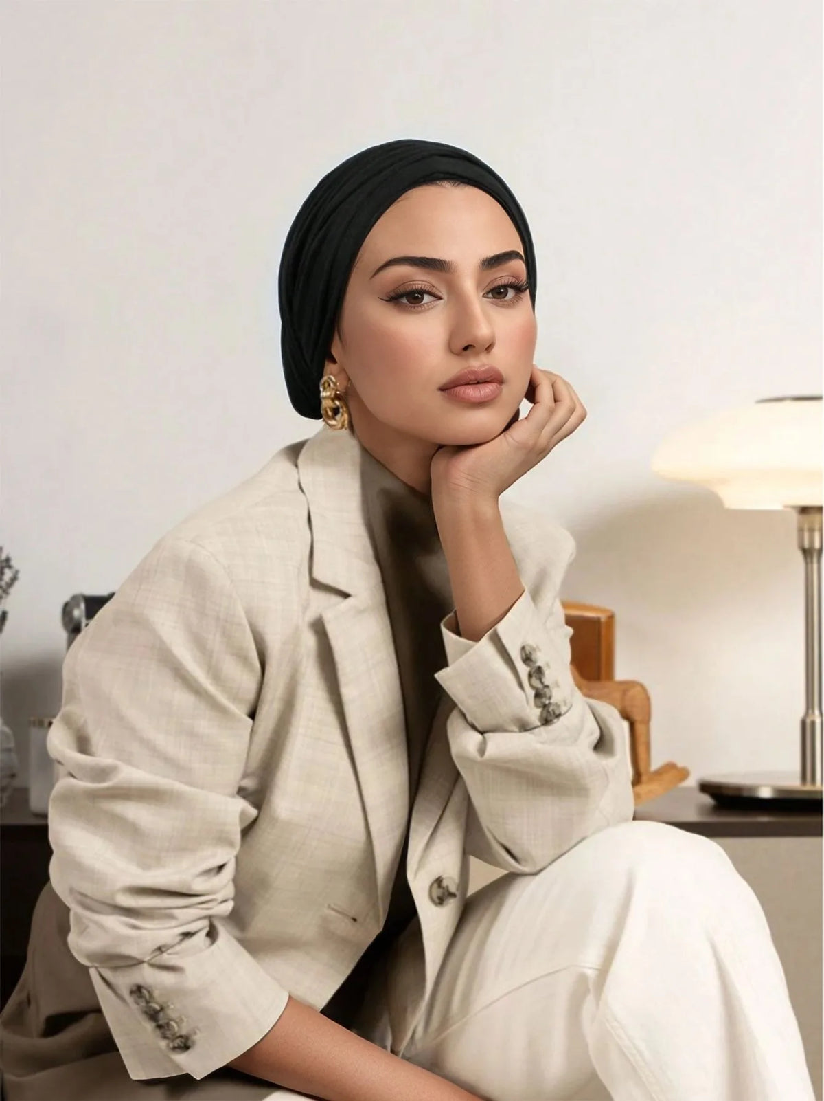 Una mujer vistiendo un blazer beige y pantalones blancos posando con la mano cerca de su rostro, mirando directamente a la cá