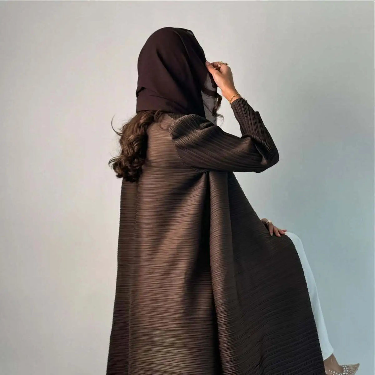 Una mujer vistiendo una cárdigan abaya plisada de color marrón oscuro, mirando por su hombro.