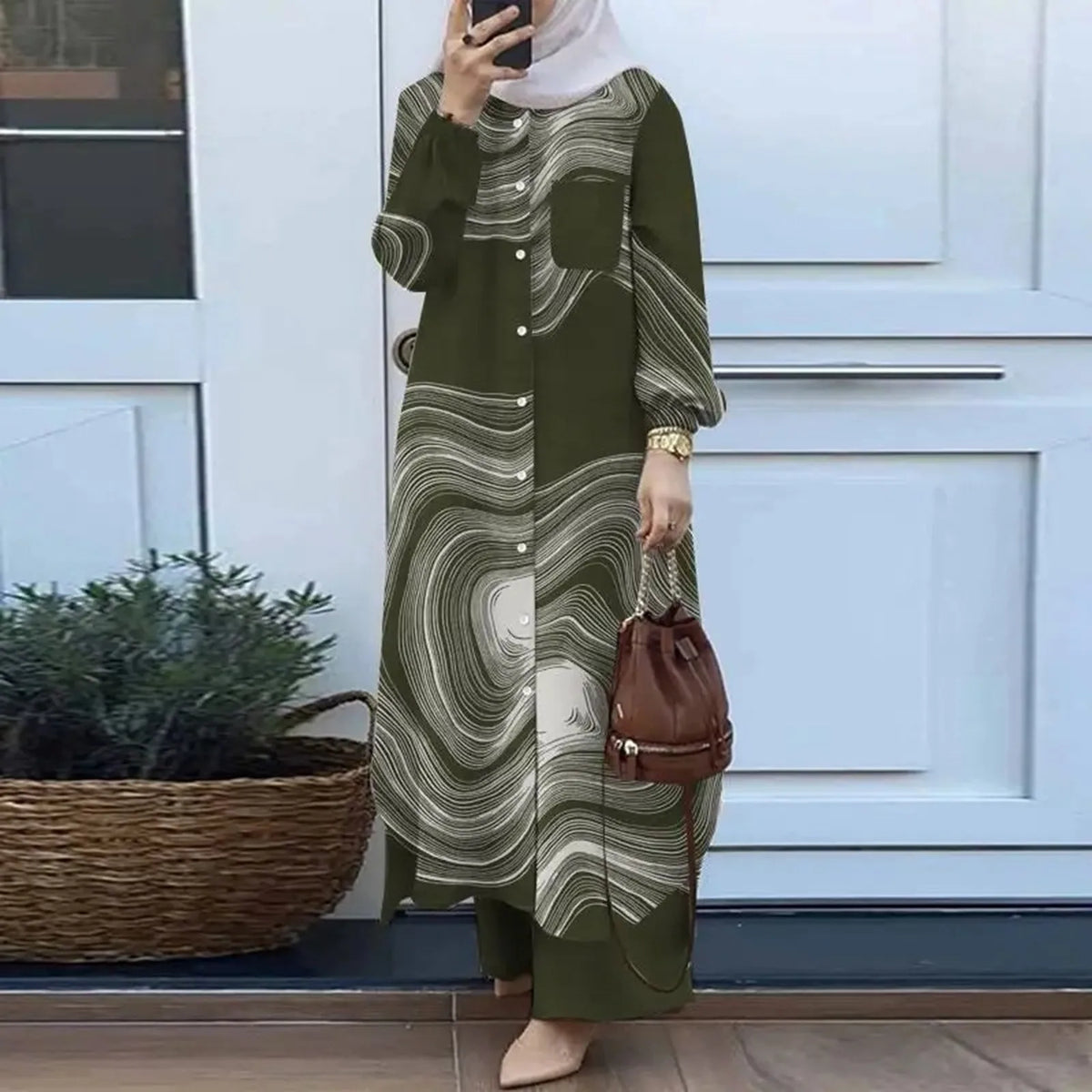 Una mujer vistiendo un conjunto abaya de color verde oliva estampado, de manga larga y un bolso marrón, frente a una puerta b