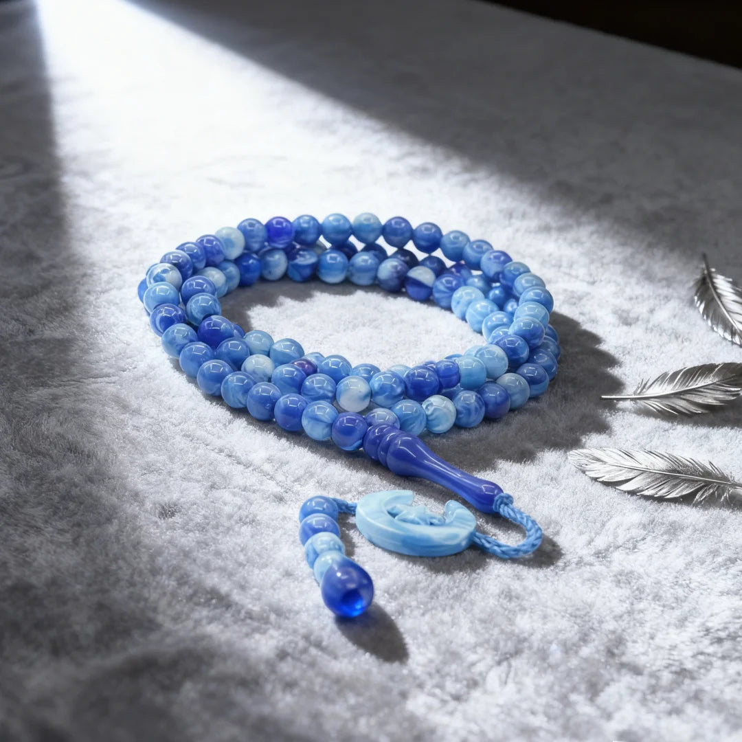 Una hebra de cuentas de vidrio azul usada como tasbih de oración con borlas de luna y estrella sobre tela blanca.