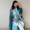 Una mujer vistiendo un cárdigan abaya plisado color verde azulado y pantalones blancos, posando con la mano cerca de su cara.
