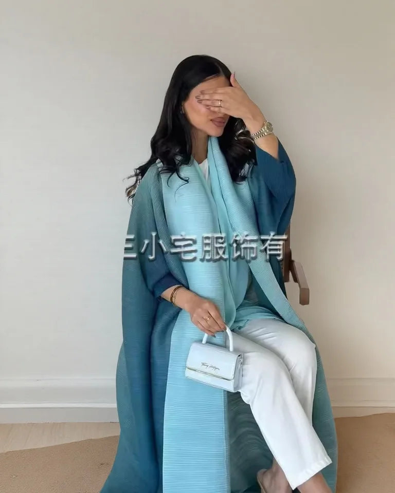 Una mujer vistiendo un cárdigan abaya plisado color verde azulado y pantalones blancos, posando con la mano cerca de su cara.