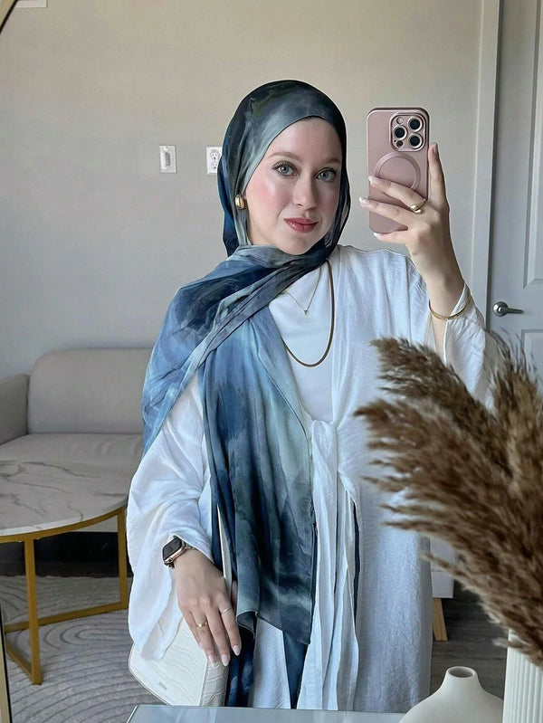 A woman wearing a blue watercolor hijab scarf and white tunic poses indoors.