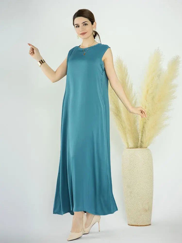 A woman models a flowing teal abaya dress against a white background, with dried yellow floral elements nearby.