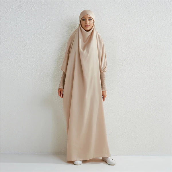 A woman wearing a beige modest abaya with smocking sleeves standing against a white wall.
