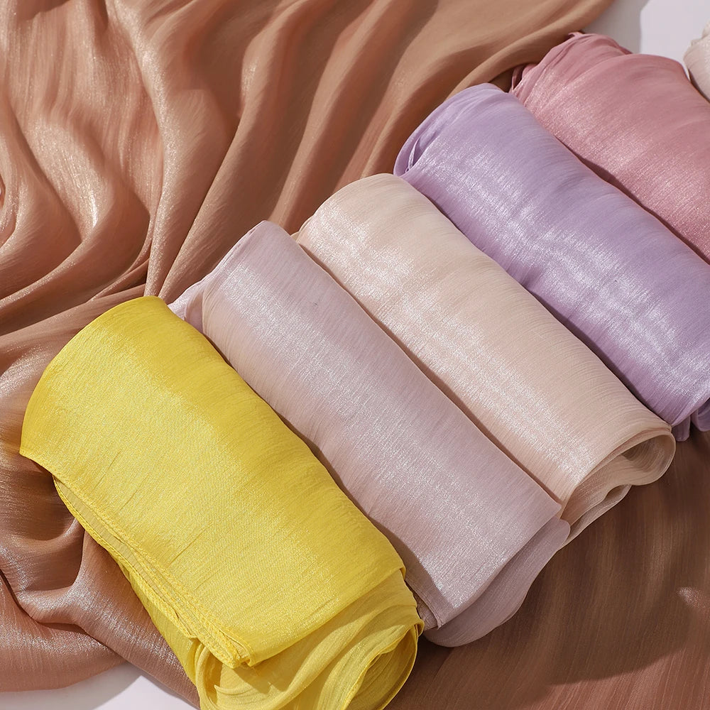 Une pile de foulards hijab en mousseline crinklée et scintillante, jaunes, beige et lilas, drapés sur un tissu