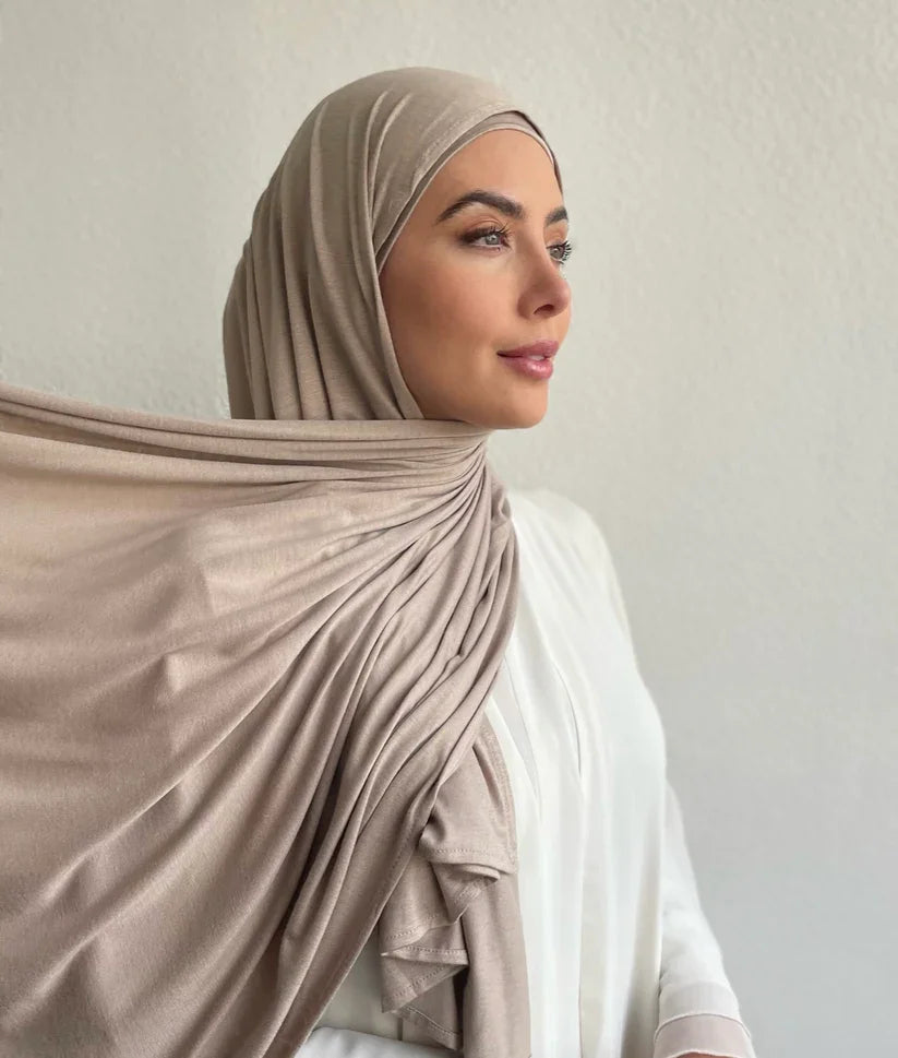 Una mujer vistiendo un hiyab y top a juego color beige, mirando hacia un lado.