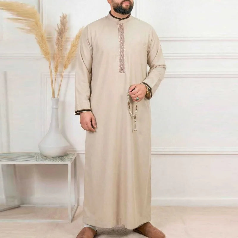 Un hombre vistiendo una jubba thobe bordada beige, bata casual, de pie en una habitación iluminada.