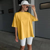 Una mujer vistiendo una camiseta básica de algodón amarilla oversize y pantalones blancos, parada frente a una puerta gris.