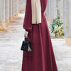 Una mujer vistiendo un vestido largo holgado color granate y hijab beige, de pie al aire libre.
