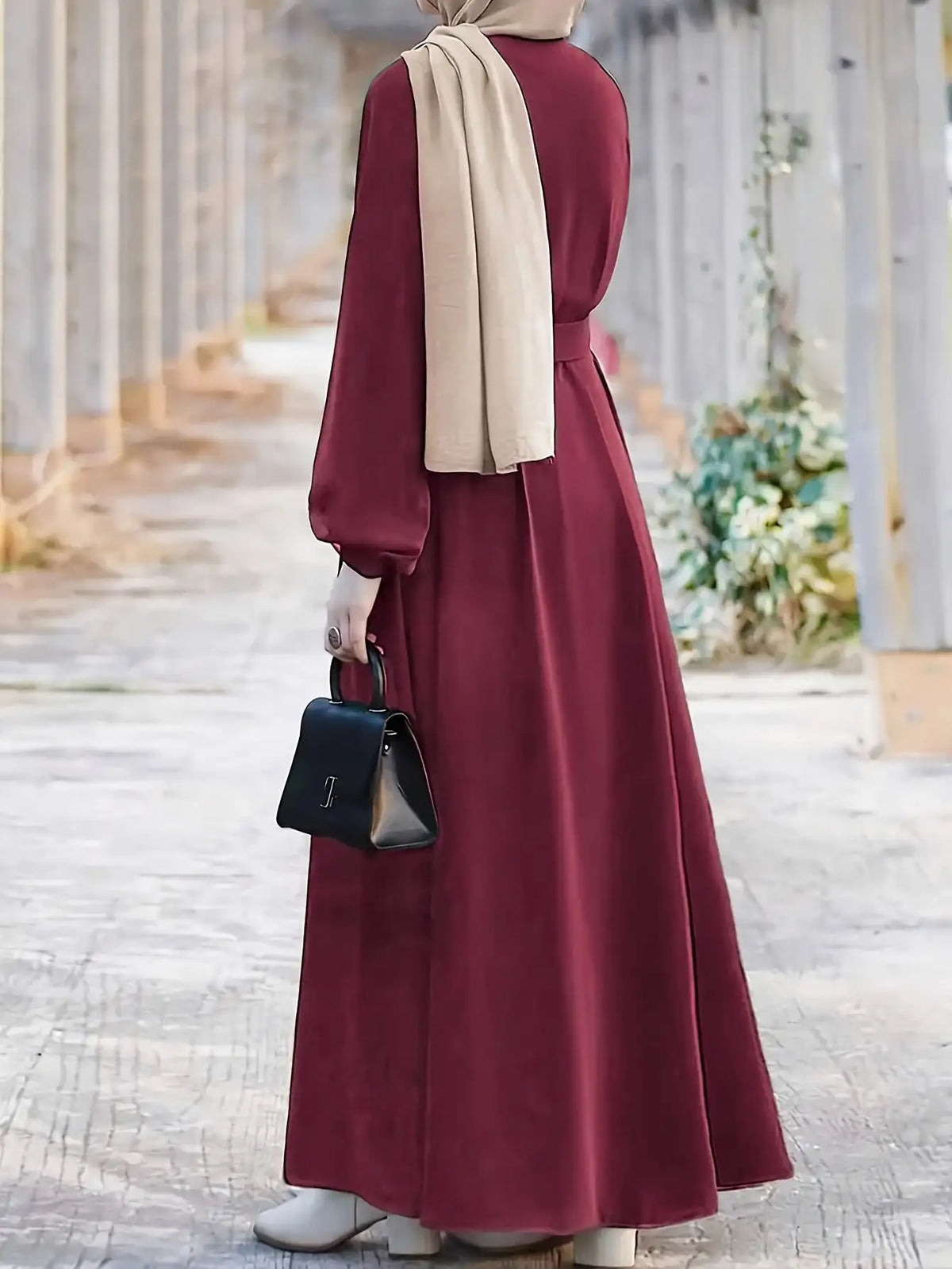 Una mujer vistiendo un vestido largo holgado color granate y hijab beige, de pie al aire libre.