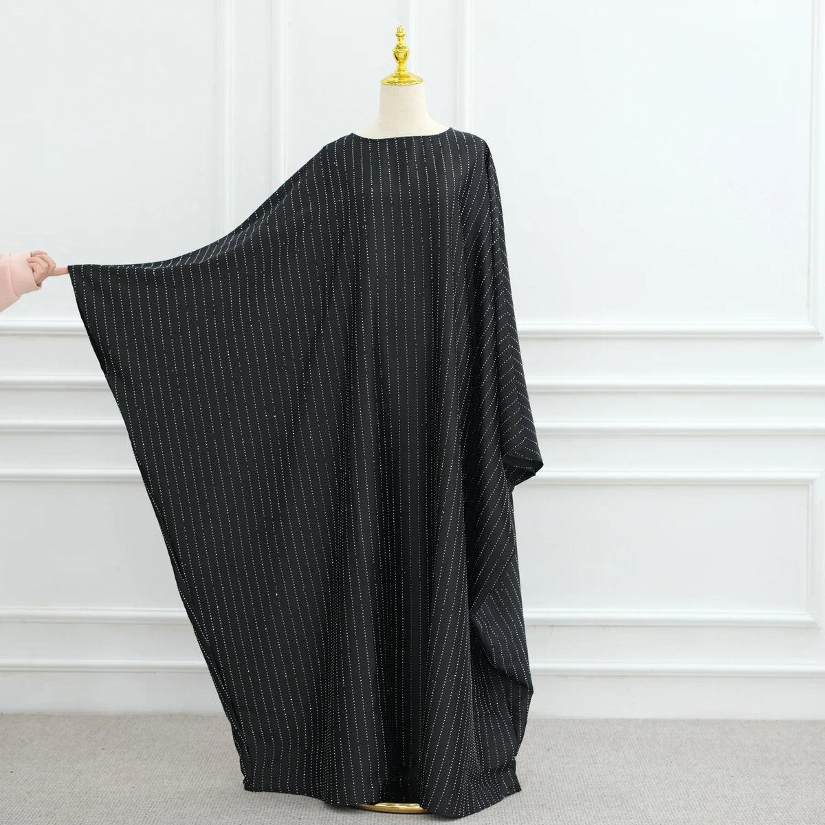 Túnica abaya de lujo con mangas de murciélago a rayas negras, exhibida en maniquí contra una pared blanca.