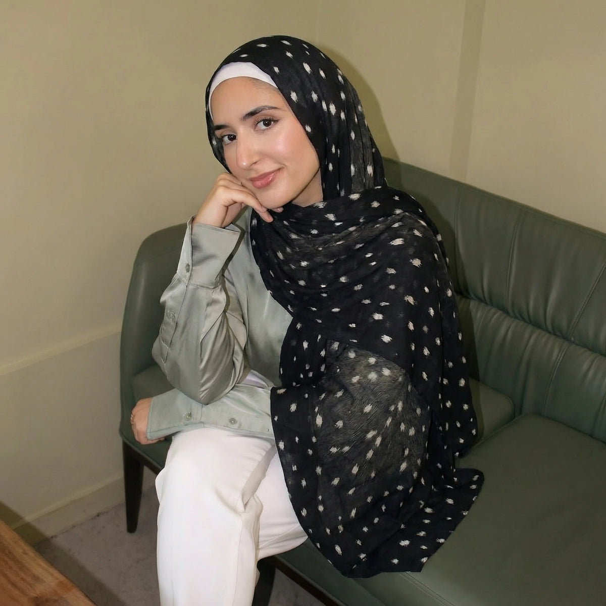 Eine Frau trägt einen schwarzen Hirschmuster-Hijabschal und ein hellgrünes Hemd und sitzt auf einem Sofa.