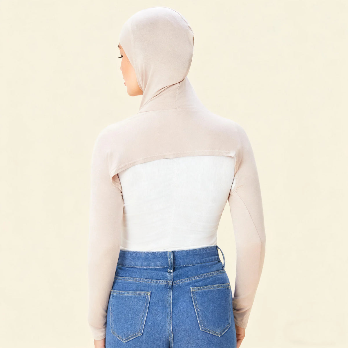 Eine Frau trägt einen beigen elastischen Modal-Jersey-Schultertop über einem weißen Oberteil und blauen Jeans.