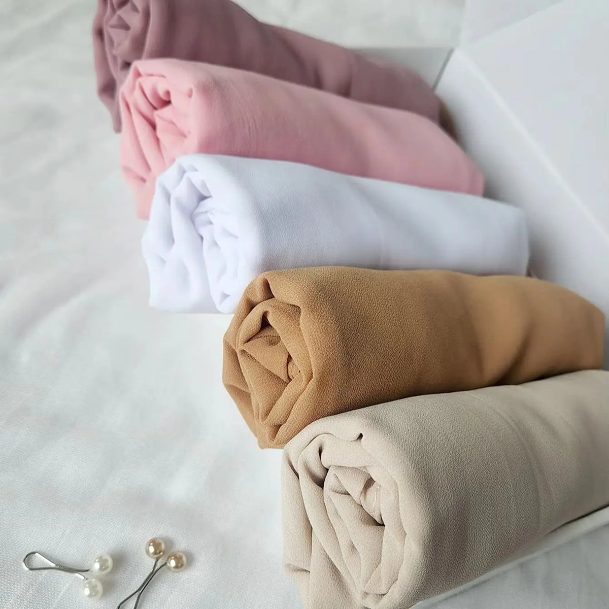 Una pila de pañuelos de chifón para hijab doblados en tonos rosa, blanco y beige sobre una superficie blanca.