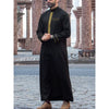 A man wearing a black embroidered thobe or jubbah with gold detailing standing outdoors.