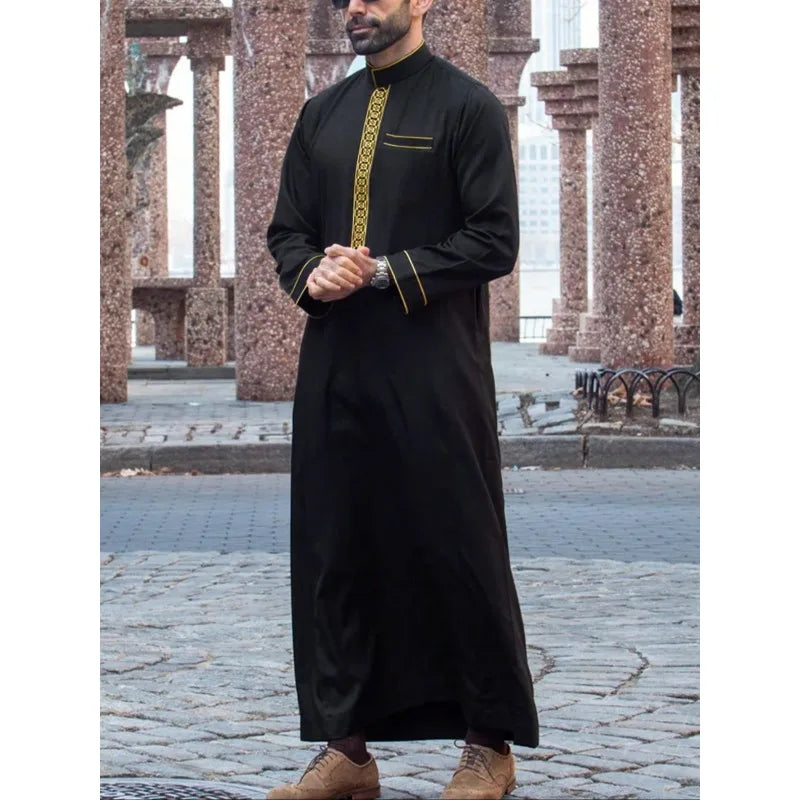 A man wearing a black embroidered thobe or jubbah with gold detailing standing outdoors.