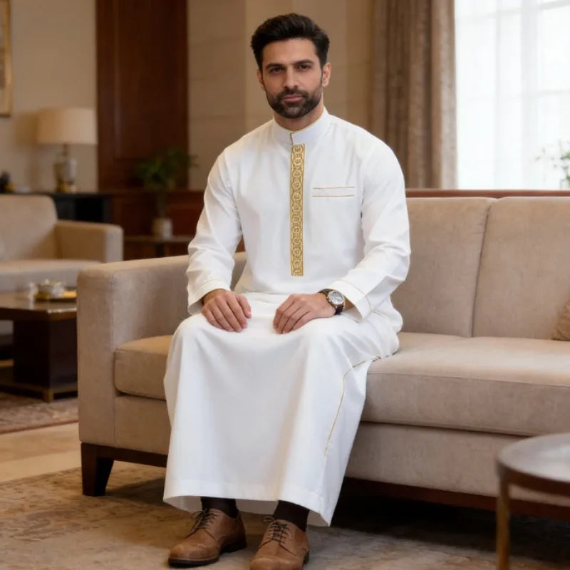 A man wearing a white embroidered thobe or jubbah sitting on a sofa in a well-lit living room.