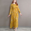 Eine Frau in einem gelb-floralen Boho-Maxikleid vor einer neutralgrauen Wand.