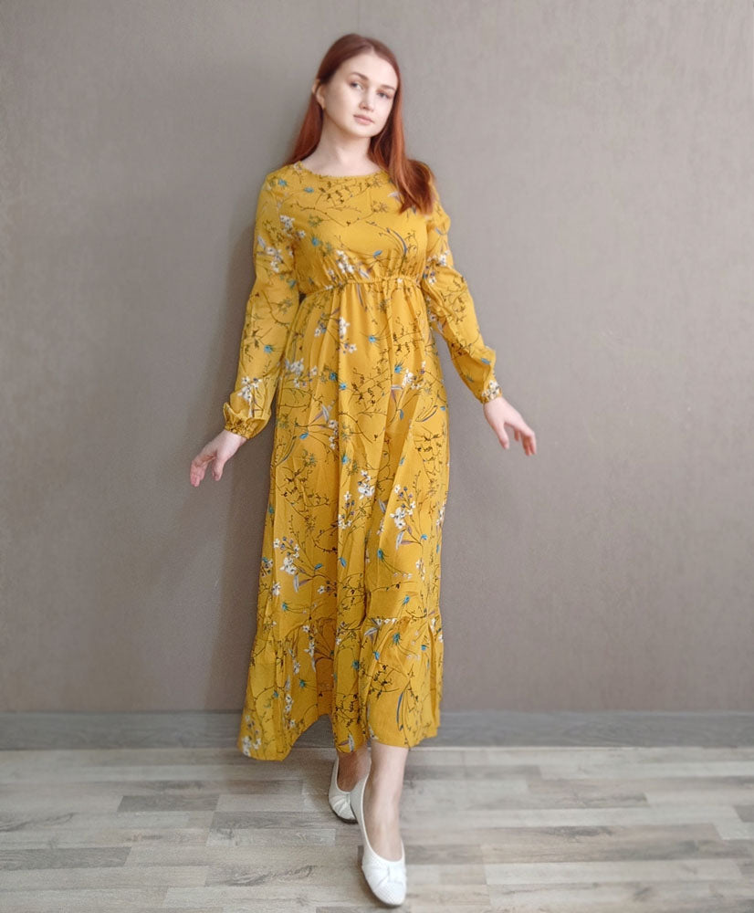 Eine Frau in einem gelb-floralen Boho-Maxikleid vor einer neutralgrauen Wand.