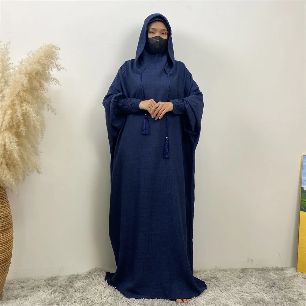Une personne portant une abaya bleu foncé avec capuche et manches batwing, debout contre un mur blanc.